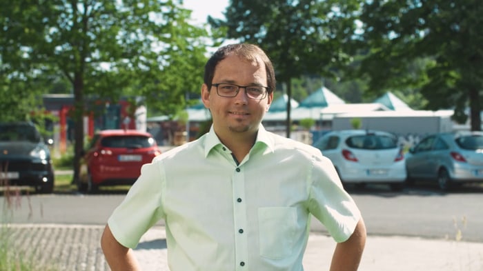 Mann mit Brille und hellgrünem Hemd steht draußen mit parkenden Autos im Hintergrund.