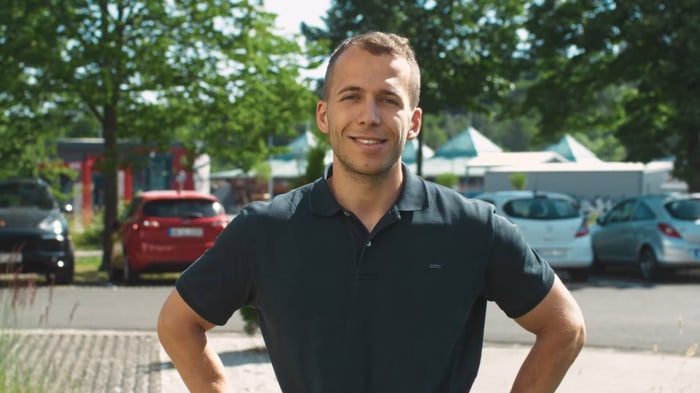 Lächelnder Mitarbeiter in dunklem Poloshirt vor dem Firmenparkplatz