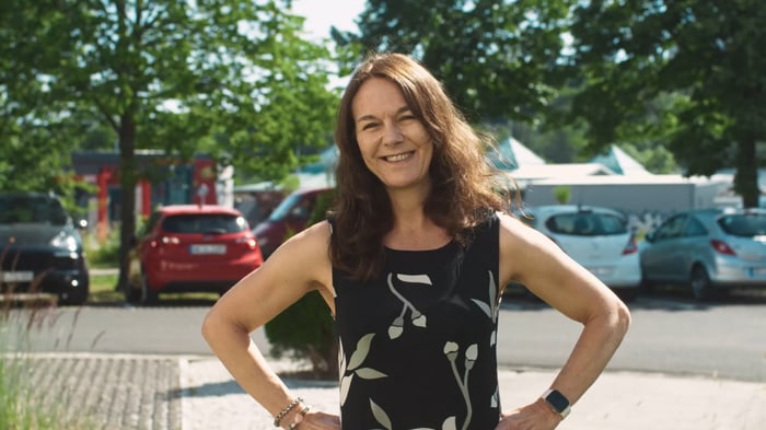 Lächelnde Mitarbeiterin in schwarzem, gemustertem Kleid vor dem Firmenparkplatz