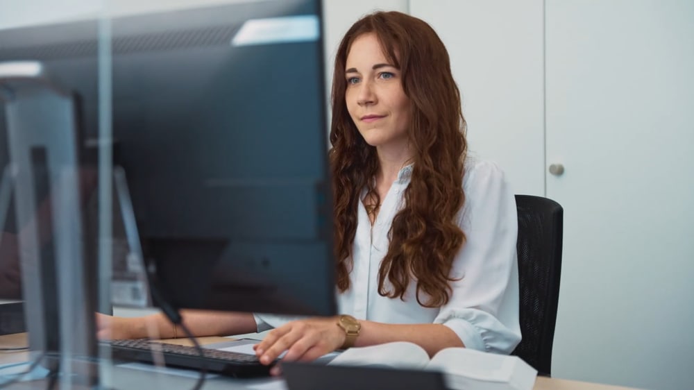 Eine junge Frau mit langen roten Haaren sitzt an einem Schreibtisch und arbeitet konzentriert am Computer.