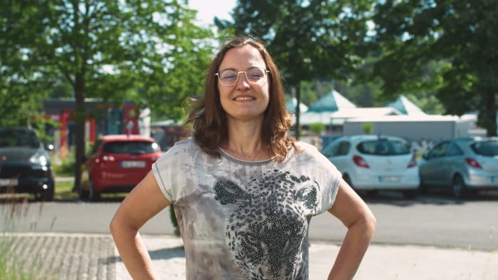 Lächelnde Mitarbeiterin mit Brille und gemustertem T-Shirt vor dem Firmenparkplatz