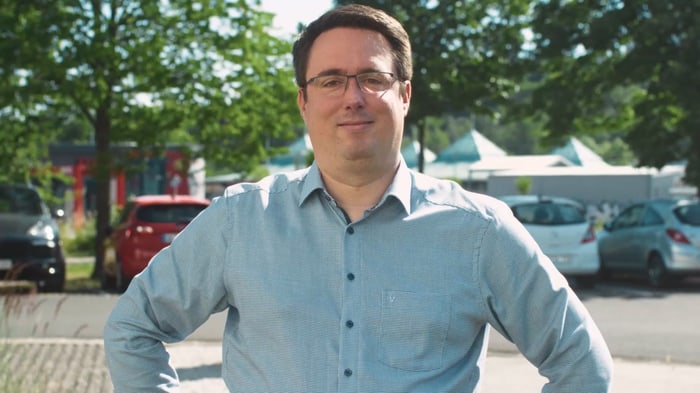 Mitarbeiter mit Brille und hellblauem Hemd vor dem Firmenparkplatz