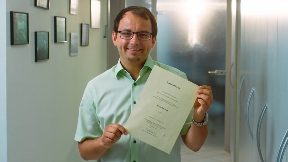 Ein Mann mit Brille und hellgrünem Hemd hält ein Dokument mit der Überschrift "Bundesrepublik" in die Kamera.